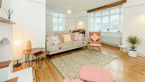 Un salon chaleureux avec un canapé, un fauteuil, une table basse et une petite table d’appoint. Il y a deux fenêtres avec des stores, un radiateur et une plante en pot.