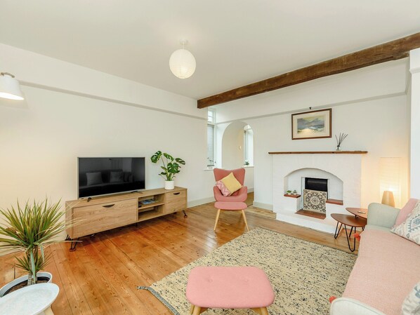 A modern living room with a flat-screen TV, wooden flooring, a pink armchair, a fireplace, and a large window. - 3 bedroom accommodation in Selsey (Selsey)