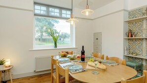 Un coin repas avec une table ronde en bois, des chaises, un vase avec des plantes vertes, une bouteille de vin et une fenêtre donnant sur des arbres.