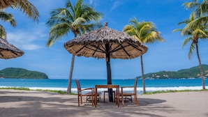 Un cadre idyllique sur la plage, avec une table et des chaises placées sous un parasol en feuilles de palmier, entourées de palmiers et offrant une vue sur l’océan.
