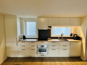 Eine moderne Küche mit weißen Schränken, einem eingebauten Backofen, einer Kaffeemaschine und zwei Fenstern mit blattförmigem Satinierglas.