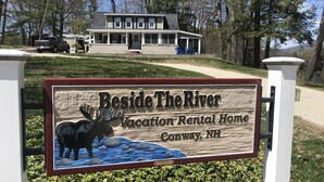 Schild für 'Beside The River Vacation Rental Home' in Conway, New Hampshire, mit einer Elch-Grafik.