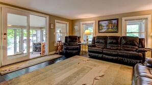 Ein geräumiges Wohnzimmer mit einer großen Ledersofa, einem Couchtisch und einem Kamin. Mehrere Fenster sorgen für natürliches Licht und einen Blick ins Freie.