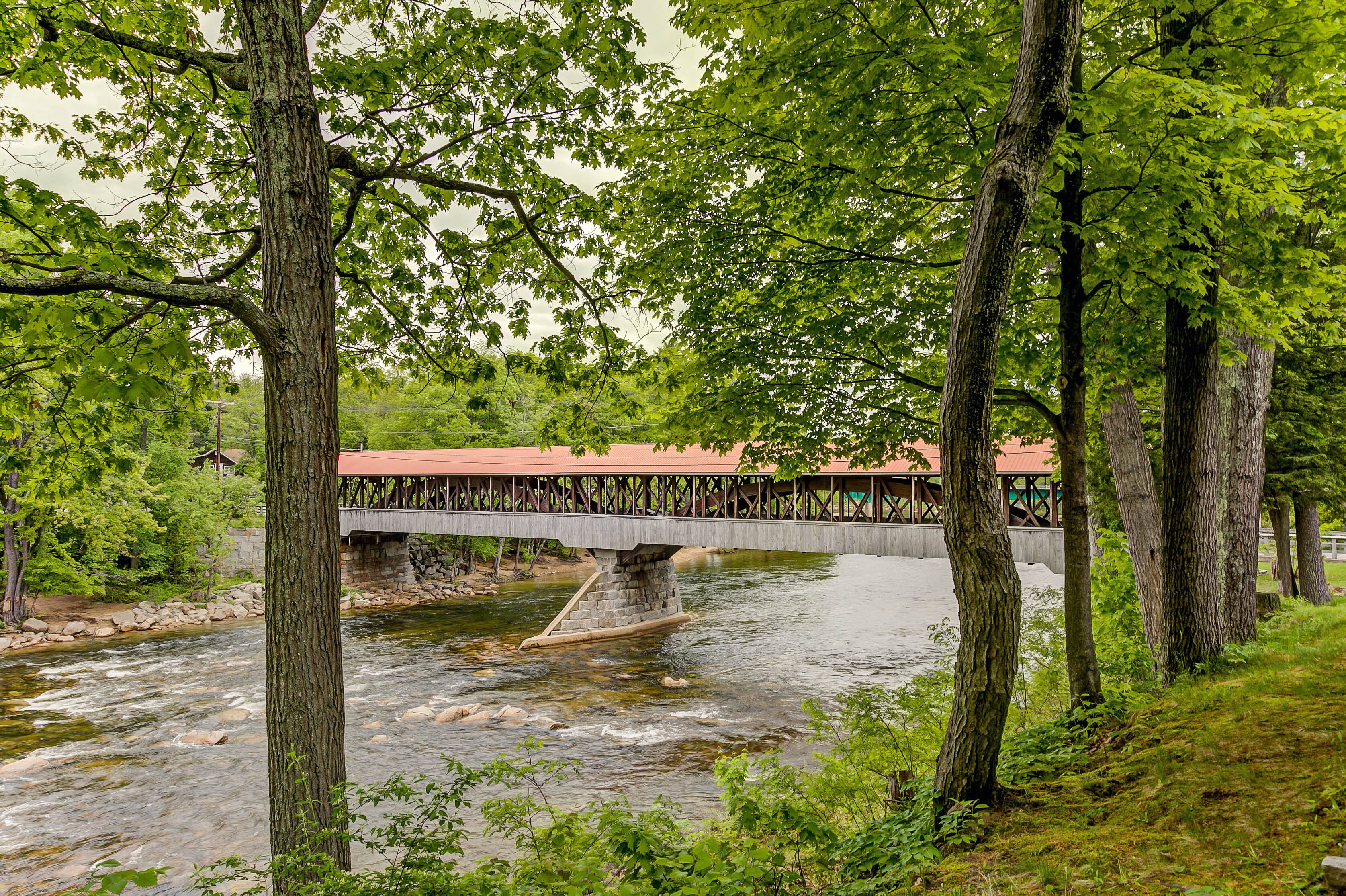 Un pont en bois enjambant une rivière, avec des piliers en pierre, entouré d’arbres et, au loin, un bâtiment doté d’un toit rouge.