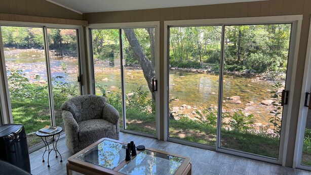 Ein Zimmer mit Glastisch, einem gemusterten Sessel und Blick auf einen Fluss durch große Schiebeglasfenster.