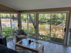 A room with a glass table, a patterned armchair, and a view of a river through large sliding glass doors. - Stay On The Saco River Next To A Covered Bridge - 2pm Check in/out (Conway)