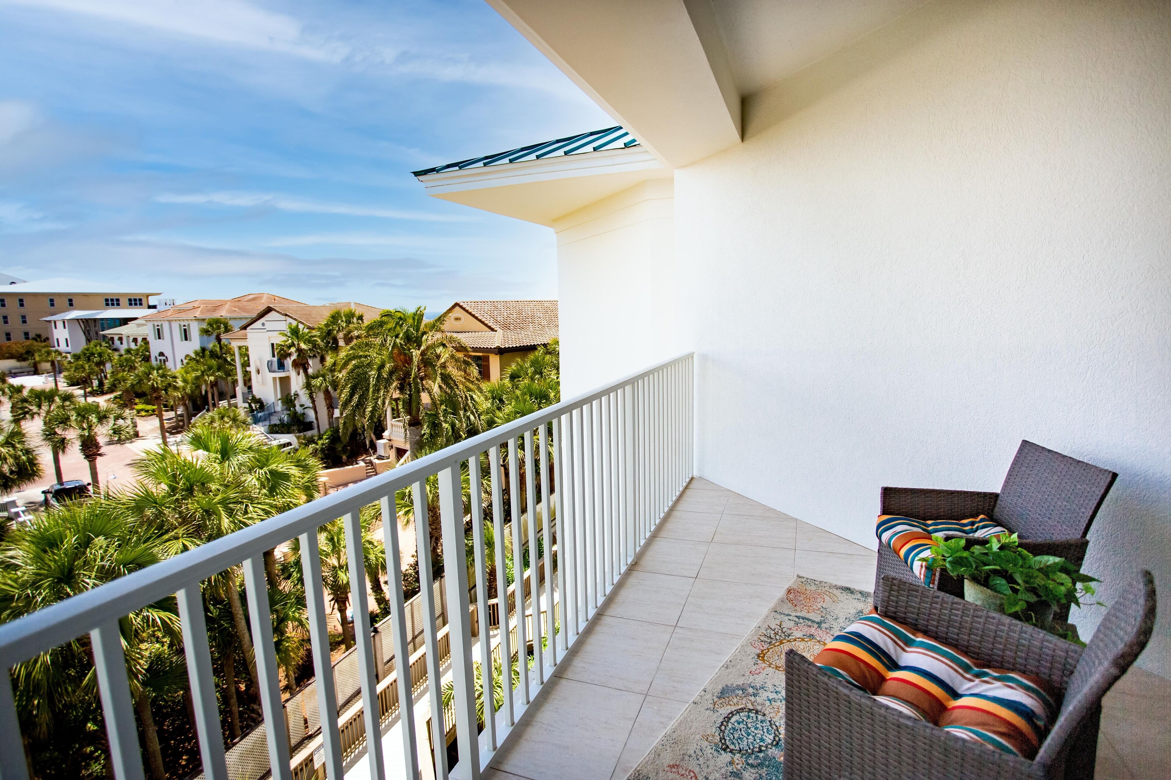 Een balkon met een rieten stoel, een plant in pot en uitzicht op een woongebied met palmbomen en huizen.