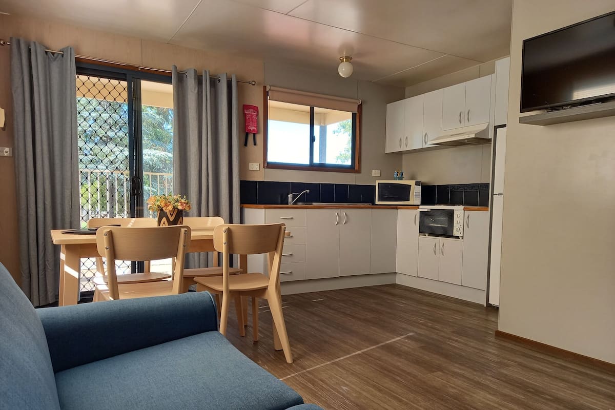 A modern kitchen with white cabinets, a microwave, and a built-in oven. There is a dining area with a table and chairs, and a blue sofa in the living area.