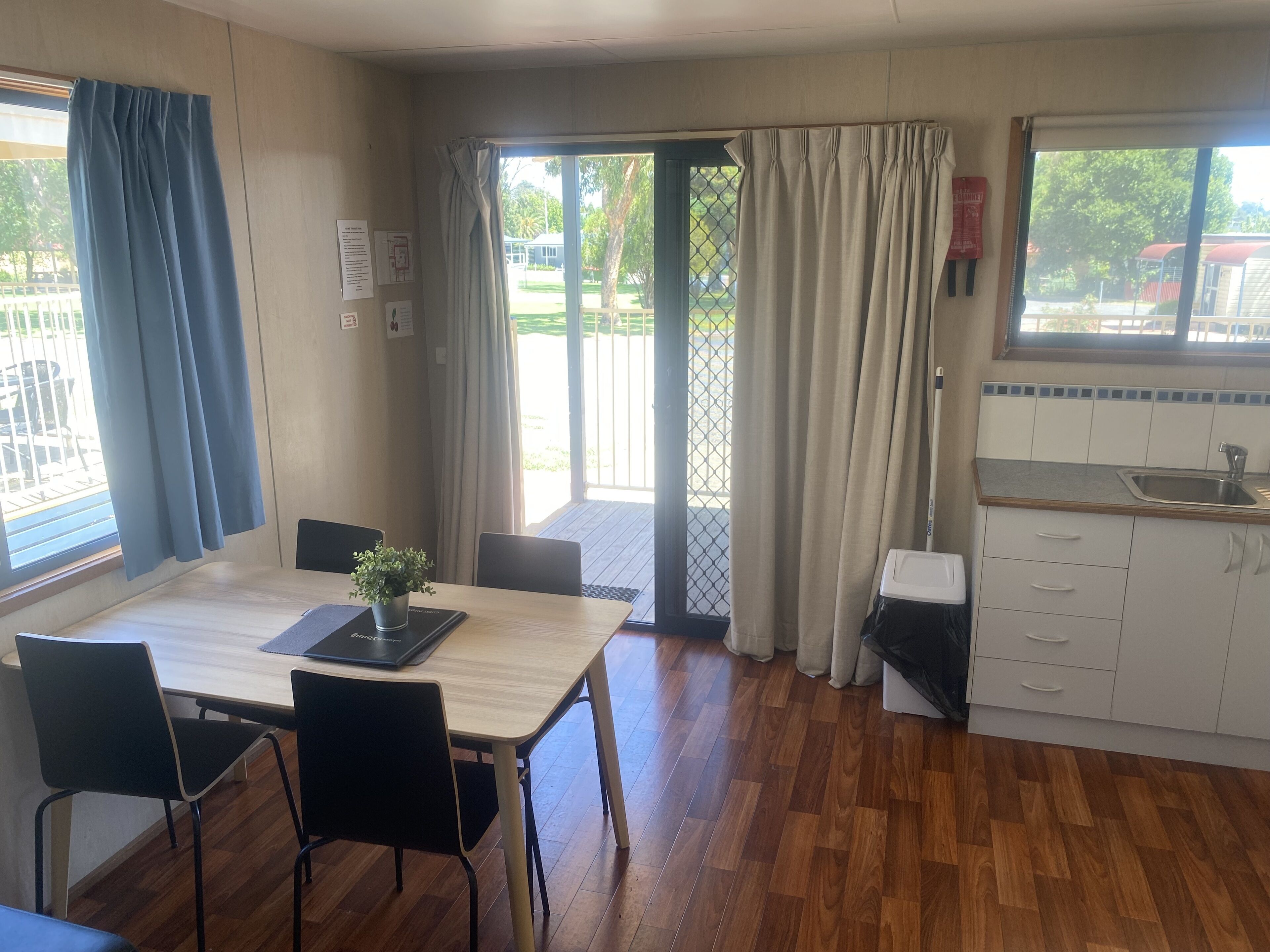 Una habitación con una mesa y sillas de madera, una pequeña kitchenette con un lavabo, y vistas a un espacio exterior a través de una puerta de vidrio.