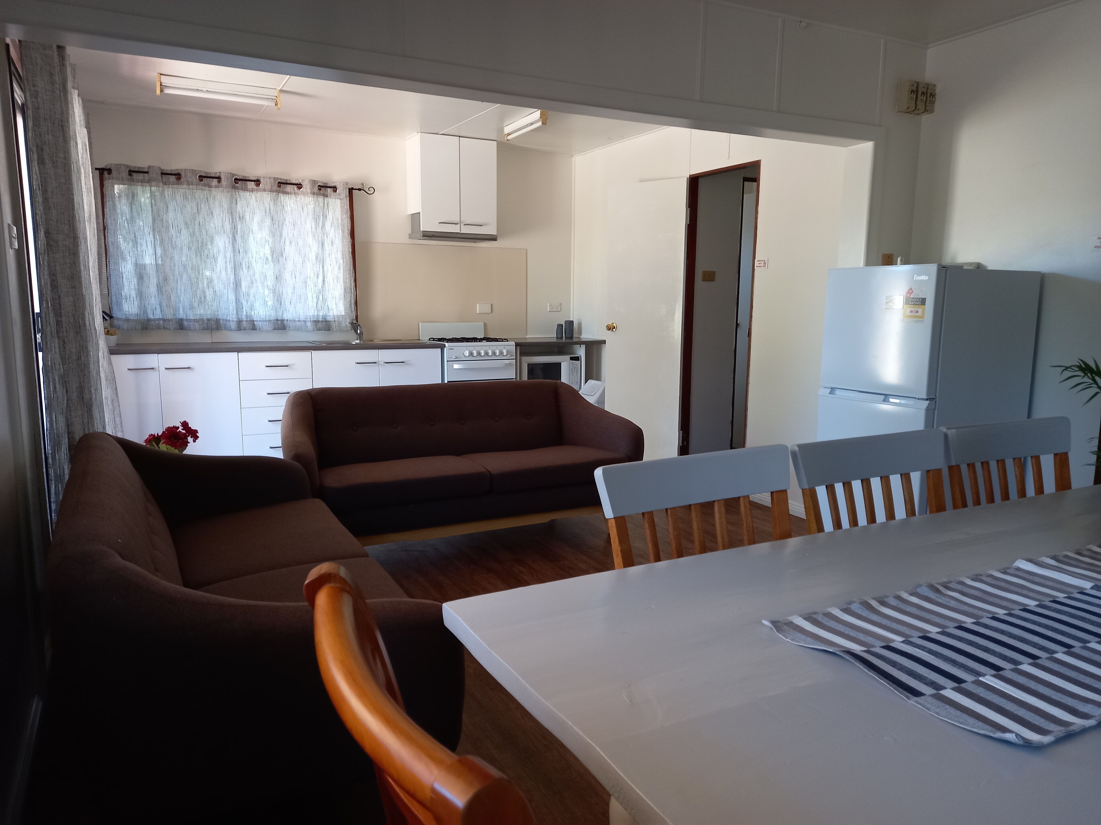 A dining area with a table, chairs, and a striped table runner. A sofa and a kitchen with cabinets and appliances are visible in the background.