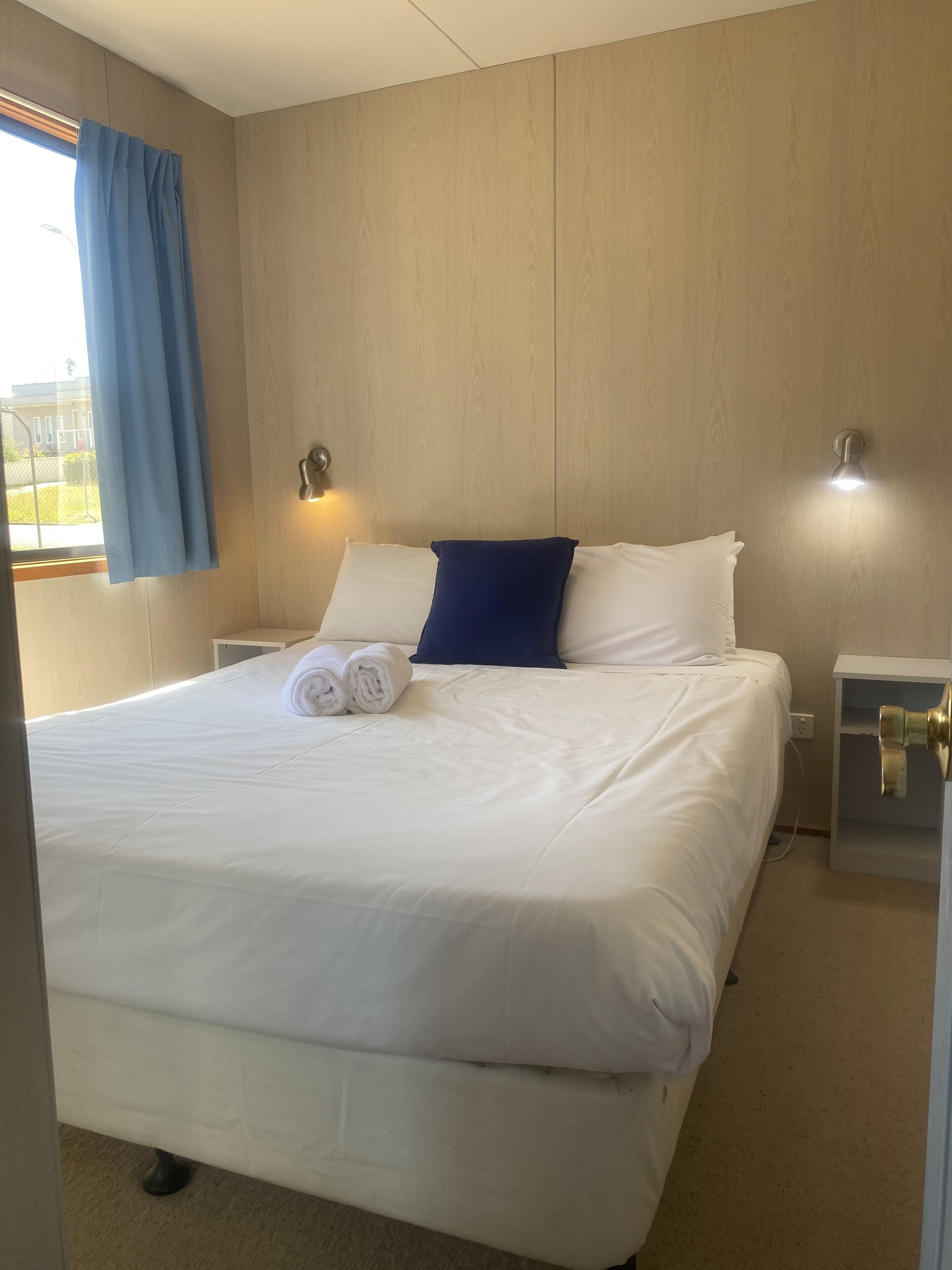 Hotellihuone, jossa on suuri sänky, kaksi yöpöytää, sinisillä verhoilla varustettu ikkuna ja seinään kiinnitetty valaisin.