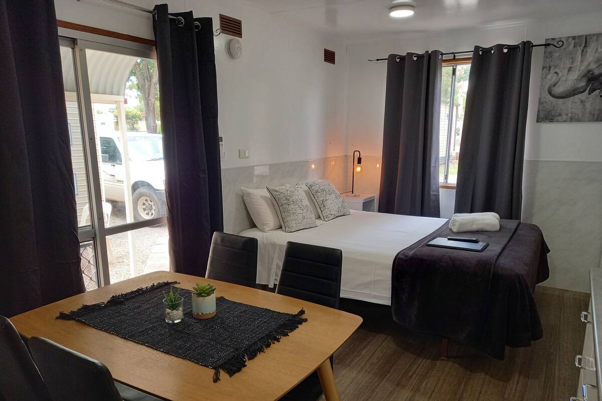 A hotel room with a bed, a small table with a plant, a bedside table with a lamp, and a view of the outside.