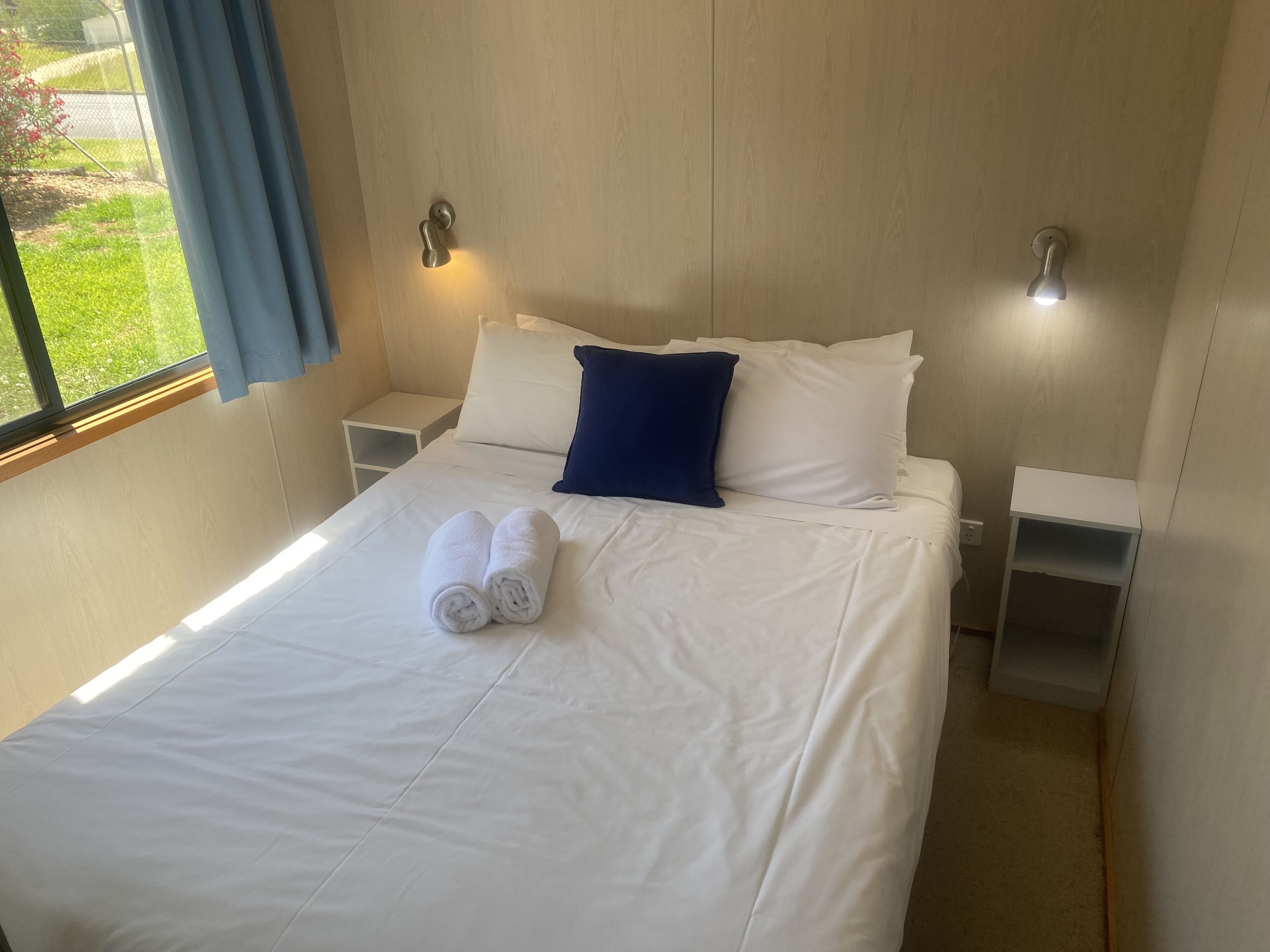 A neatly made bed with white linens and two rolled towels, a blue pillow, and two bedside tables with wall-mounted lamps.