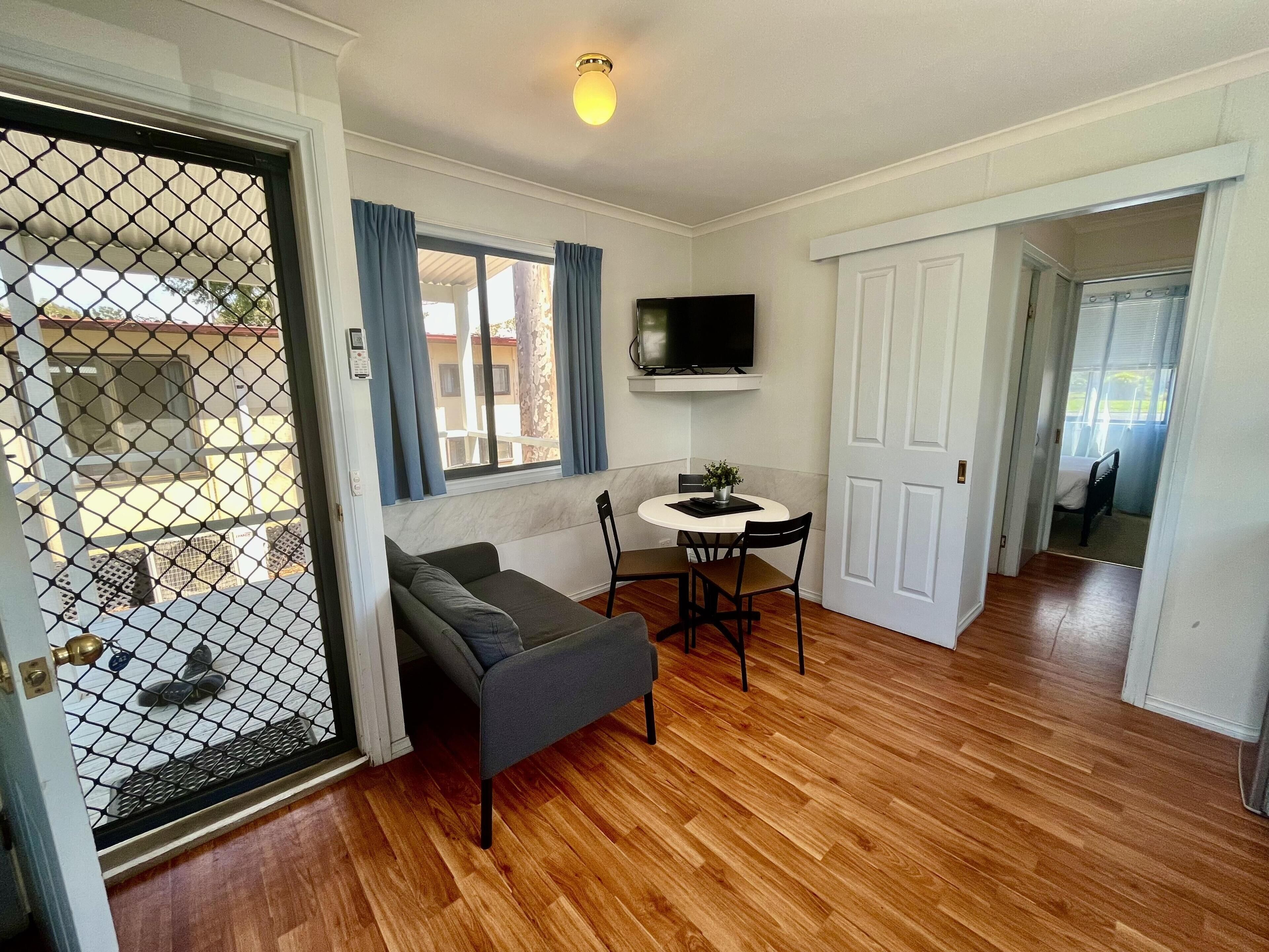 A room with a sofa, a dining table with chairs, a TV mounted on the wall, and a door leading to another room.