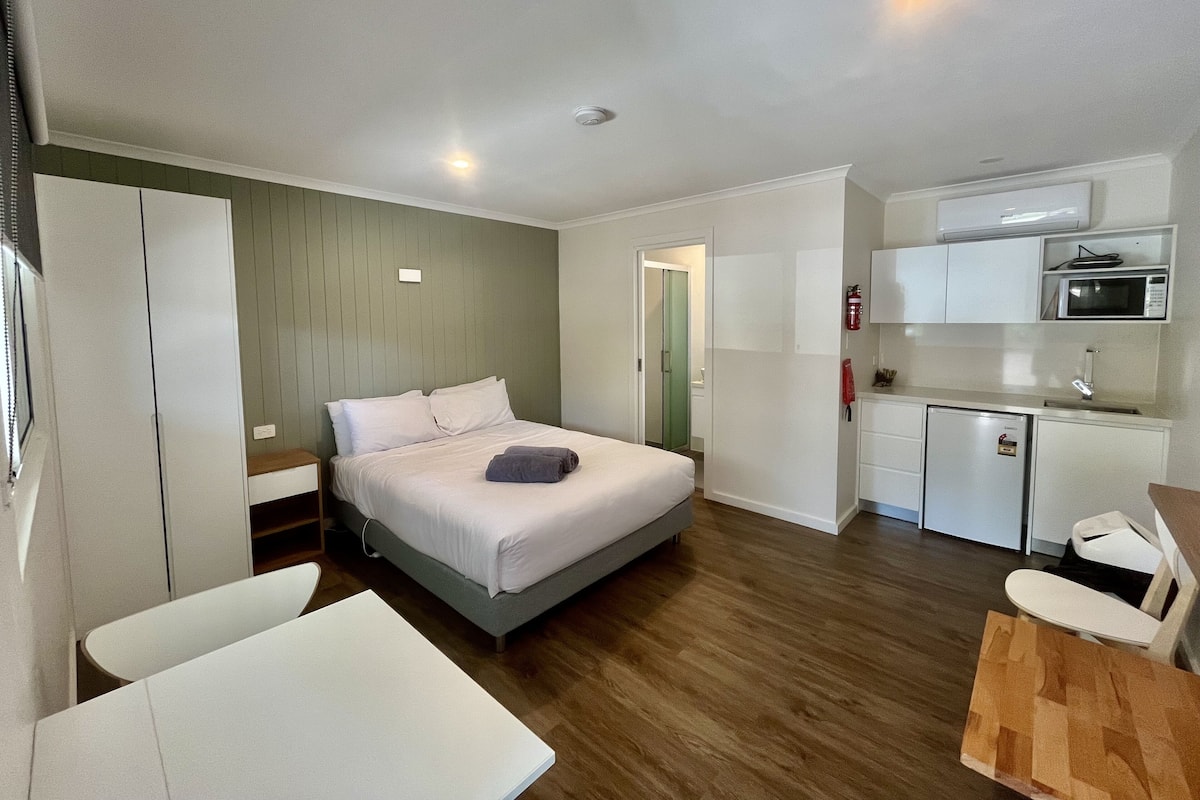 A compact hotel room with a bed, a small kitchenette, a dining area, and a bathroom.