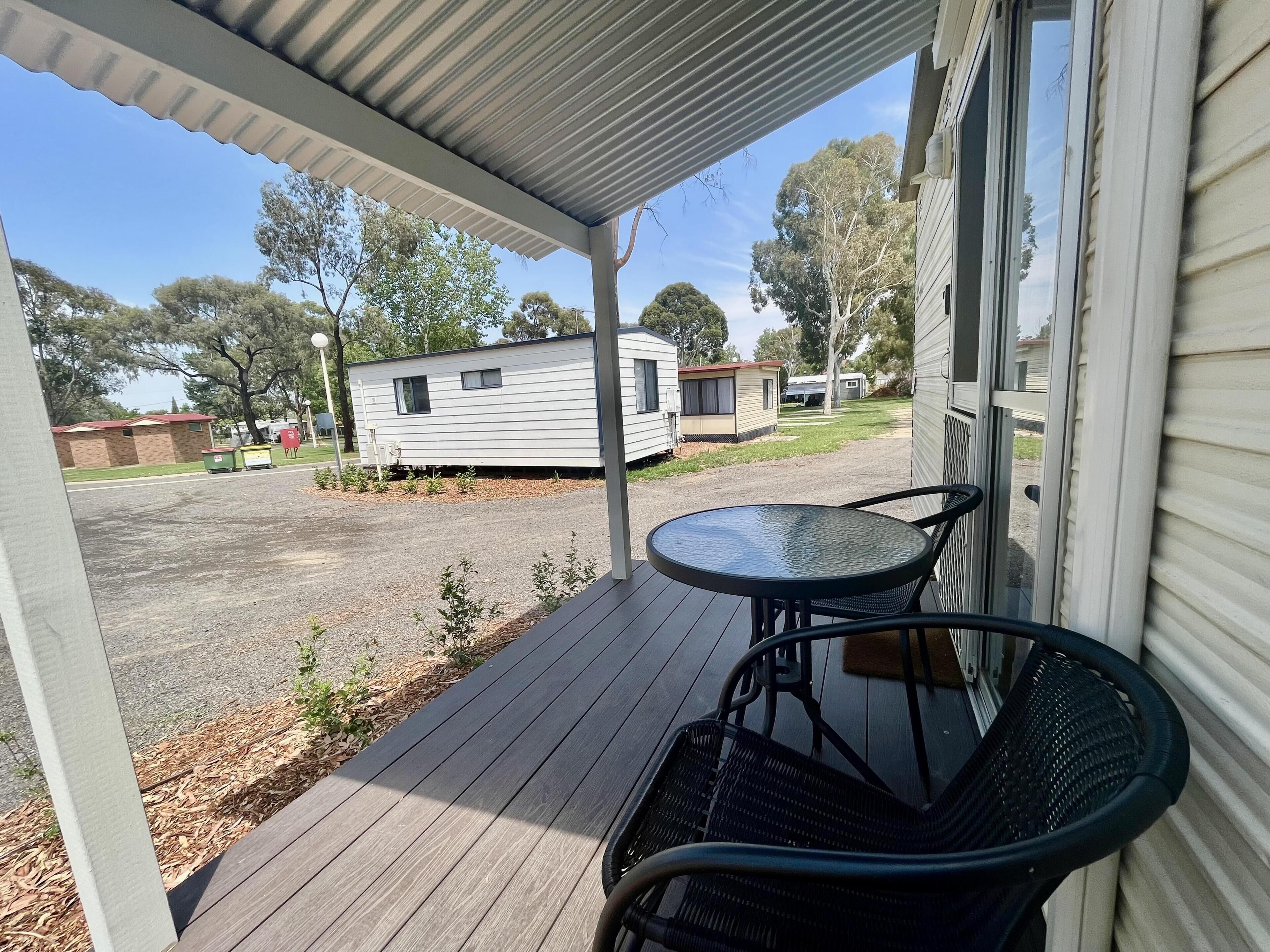 A covered porch with a table and chairs, a view of a parking lot, and mobile homes in the background.