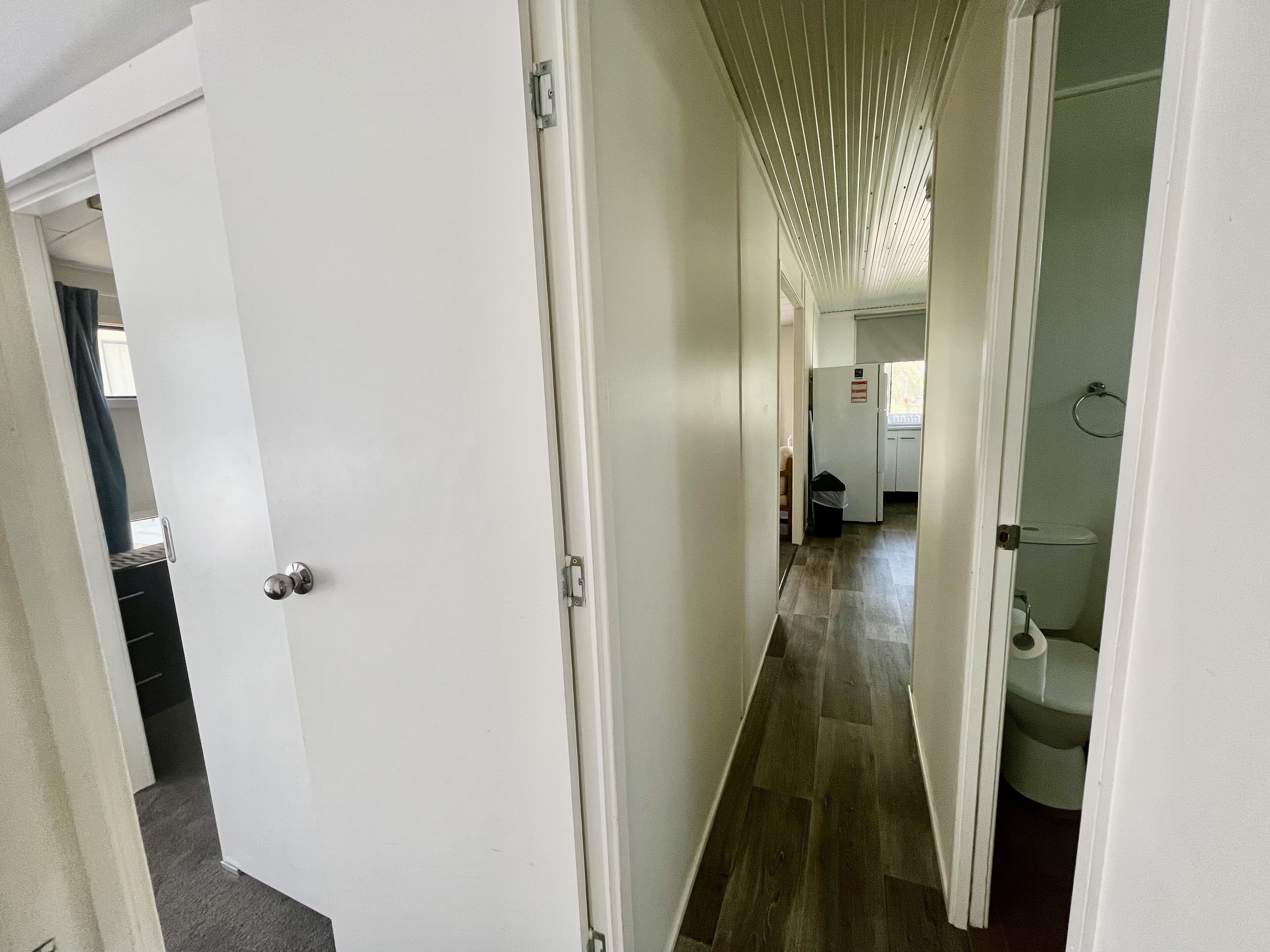 Een smalle gang met witte muren, een houten vloer en een plafond met lineaire verlichting. Links staat een deur open, die een kamer onthult met een bed en een raam.