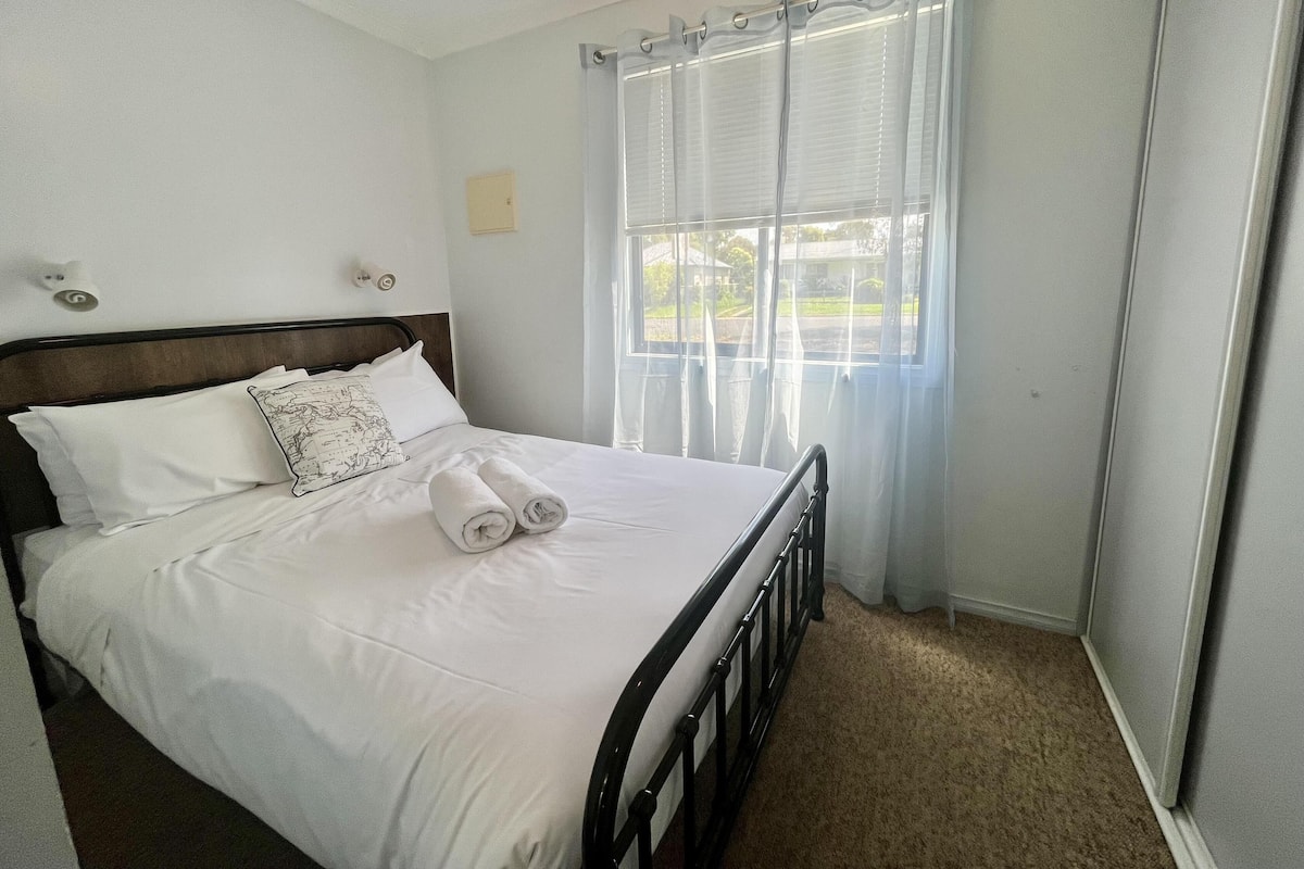 A neatly made bed with white linens and rolled towels, a wooden headboard, a window with sheer curtains, and a carpeted floor.