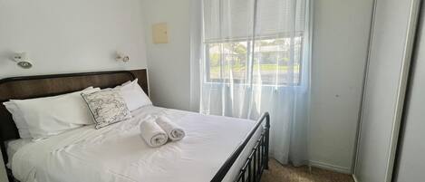 A neatly made bed with white linens and rolled towels, a wooden headboard, a window with sheer curtains, and a carpeted floor.
