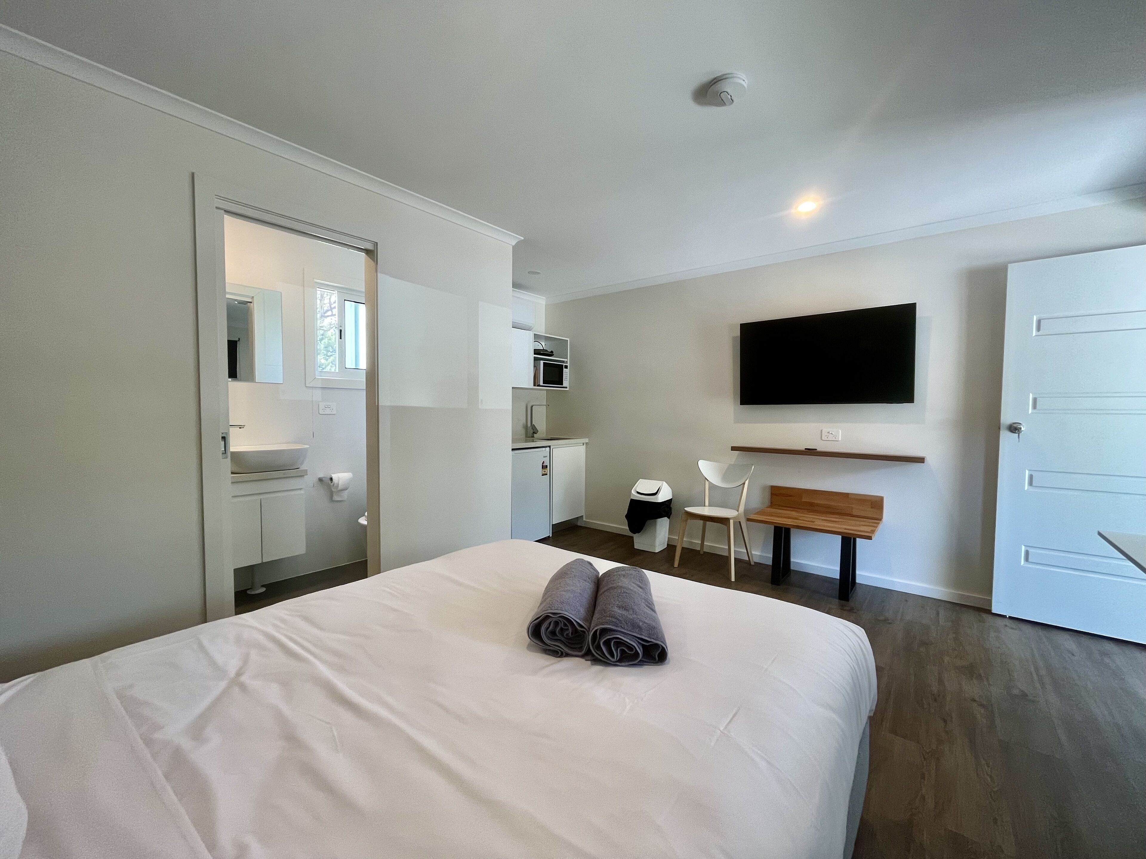 A modern hotel room with a large bed, a TV mounted on the wall, a small kitchenette, and a bathroom visible through an open door.