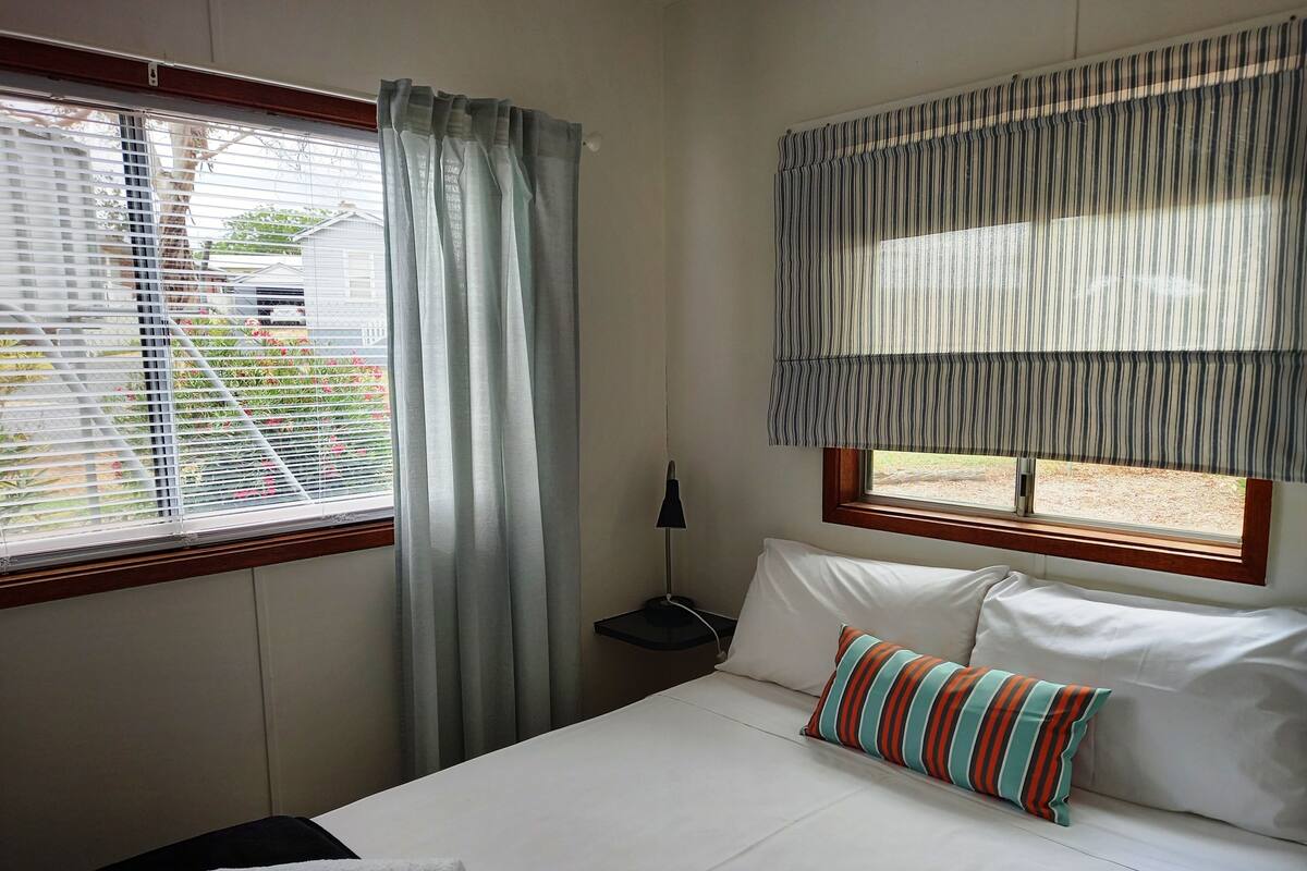 A hotel room with a bed, a window with blinds, a nightstand, and a lamp.