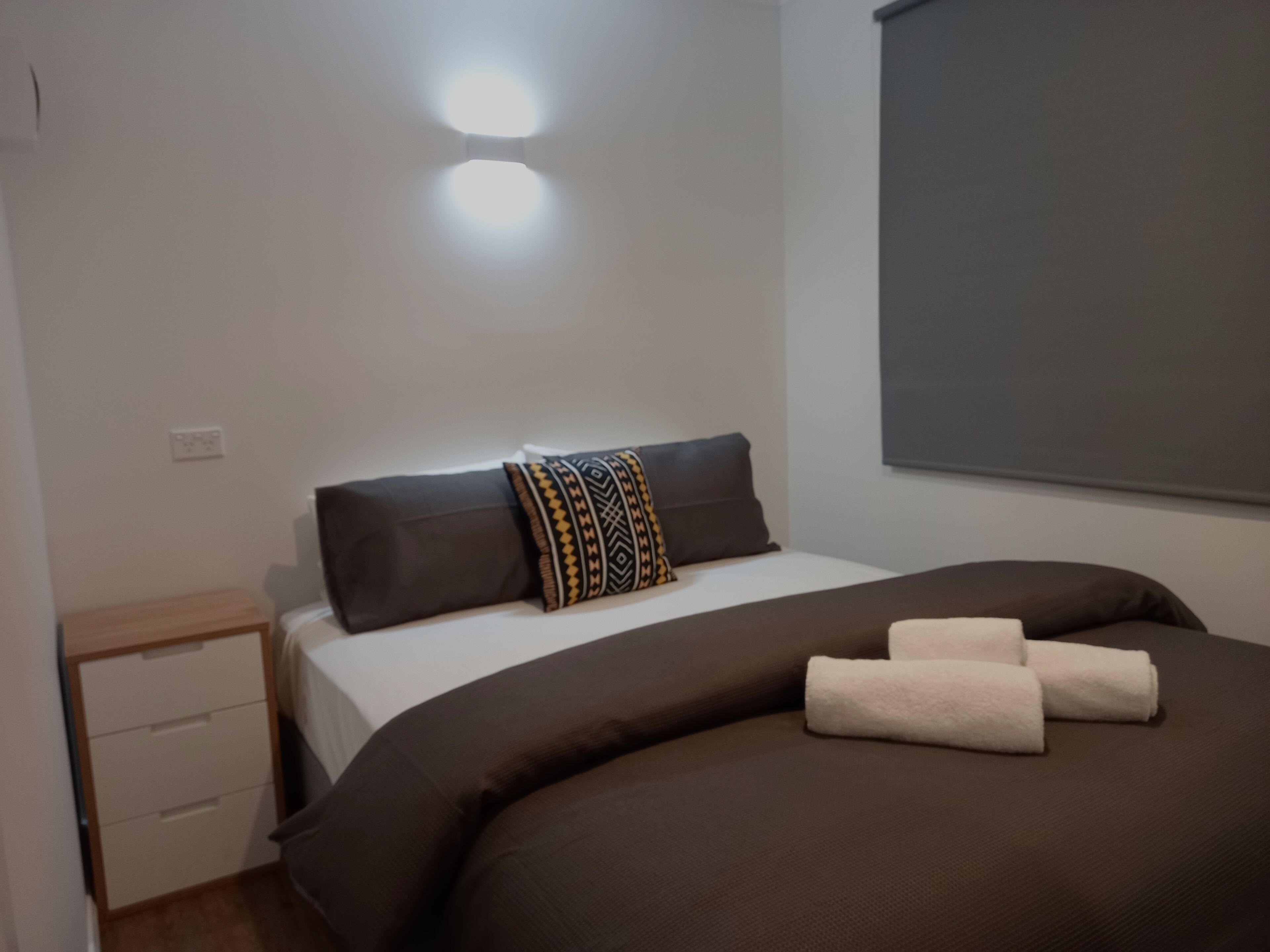 A neatly made bed with a patterned pillow, a bedside table with a drawer, and a wall-mounted light fixture.