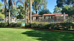 A well-maintained grassy area with a pool, surrounded by palm trees and a fenced building with a red roof. - Young Tourist Park (Young)
