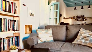 Un salon moderne avec un canapé gris, une table basse en verre et un ordinateur portable. On y trouve des étagères remplies de livres et une plante, un cactus.