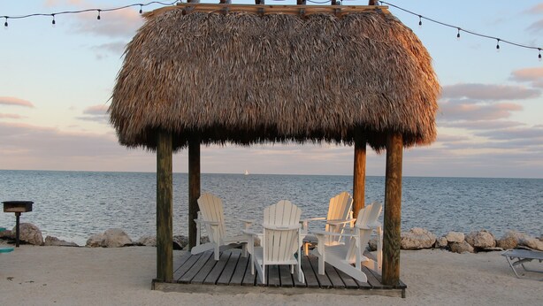 A beachside gazebo with a thatched roof, wooden pillars, and white Adirondack chairs.
