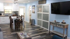 A modern kitchen with white cabinets, a dining area with wooden chairs, a TV mounted on the wall, and a console table with decorative items.