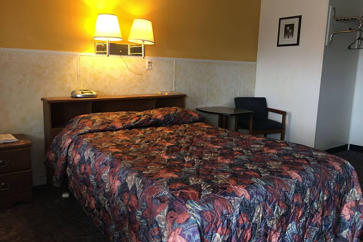 Una habitación de hotel con cama, mesita de noche, lámpara, silla y una pequeña mesa.