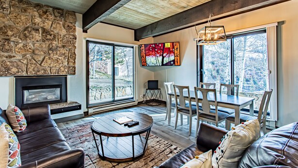 A modern living room with a stone fireplace, large windows, a dining area, and a cozy seating arrangement. - Ski-In/Ski-Out Condo | Storm Meadows Club A! (Steamboat Springs)