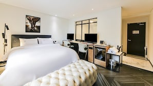 Una habitación de hotel moderna con una cama grande, un escritorio con dos monitores, un sillón cómodo y un espejo amplio.