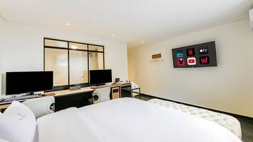 Una habitación de hotel con una cama, un escritorio con un ordenador, dos monitores, un televisor y una pantalla mural que muestra logotipos de canales de televisión.