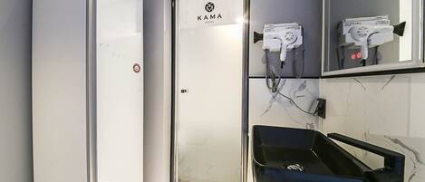 Un baño de hotel con un refrigerador blanco, un espejo con el letrero KAMA HOTEL y un lavamanos negro.