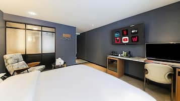 A modern hotel room with a bed, a desk, a chair, a TV displaying streaming service logos, and a large window with frosted glass.