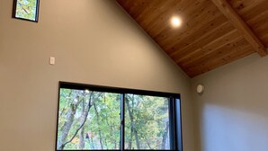 Une pièce dotée d’un sol recouvert de tatamis, d’une grande fenêtre offrant une vue sur les arbres, de poutres en bois au plafond et d’un éclairage encastré.