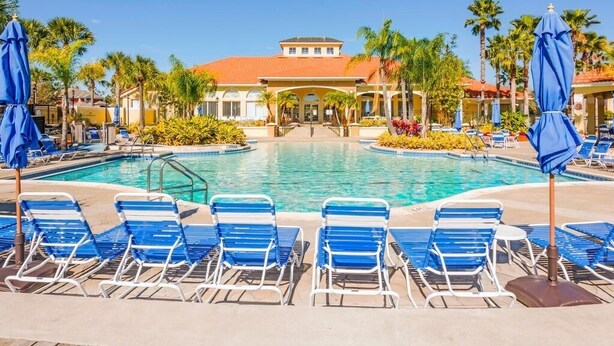 A pool area with blue lounge chairs and umbrellas, surrounded by palm trees and a building with a red roof.