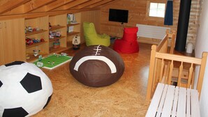 Un acogedor cuarto ático con vigas de madera, un televisor, una estantería con juguetes, un sillón de peluche rojo y una pelota de fútbol.