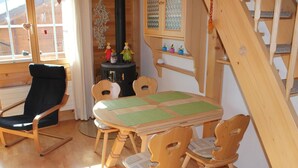 Una habitación acogedora con una mesa y sillas de madera, un sillón mecedora negro, una mesita con adornos y una ventana con cortinas.