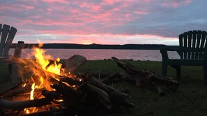 Un feu de camp avec deux chaises Adirondack placées face à un lac, au coucher du soleil.