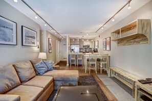 Un salon moderne avec un canapé d’angle, un coin repas avec une table et des chaises, et une cuisine en arrière-plan.