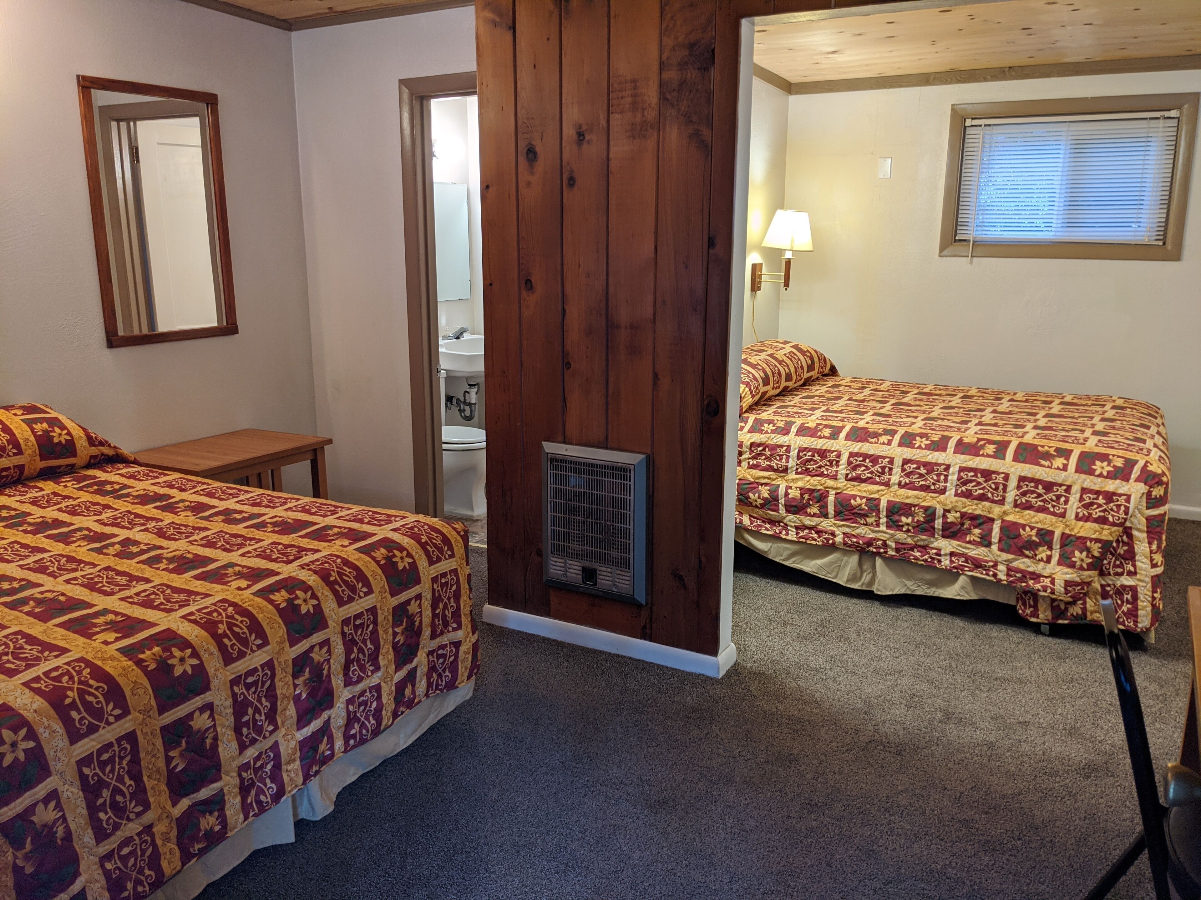 A bedroom with two beds, a wooden wall, a mirror, and a window with blinds.