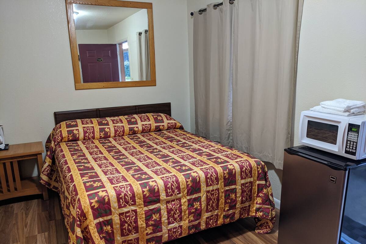 A room with a bed, a wooden nightstand, a mirror, a microwave, and a refrigerator.
