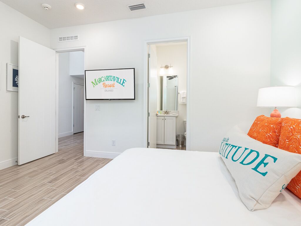 Une chambre d’hôtel avec un lit, un oreiller orné du mot « ATTITUDE », un panneau encadré accroché au mur, et une salle de bain visible à travers une porte ouverte.