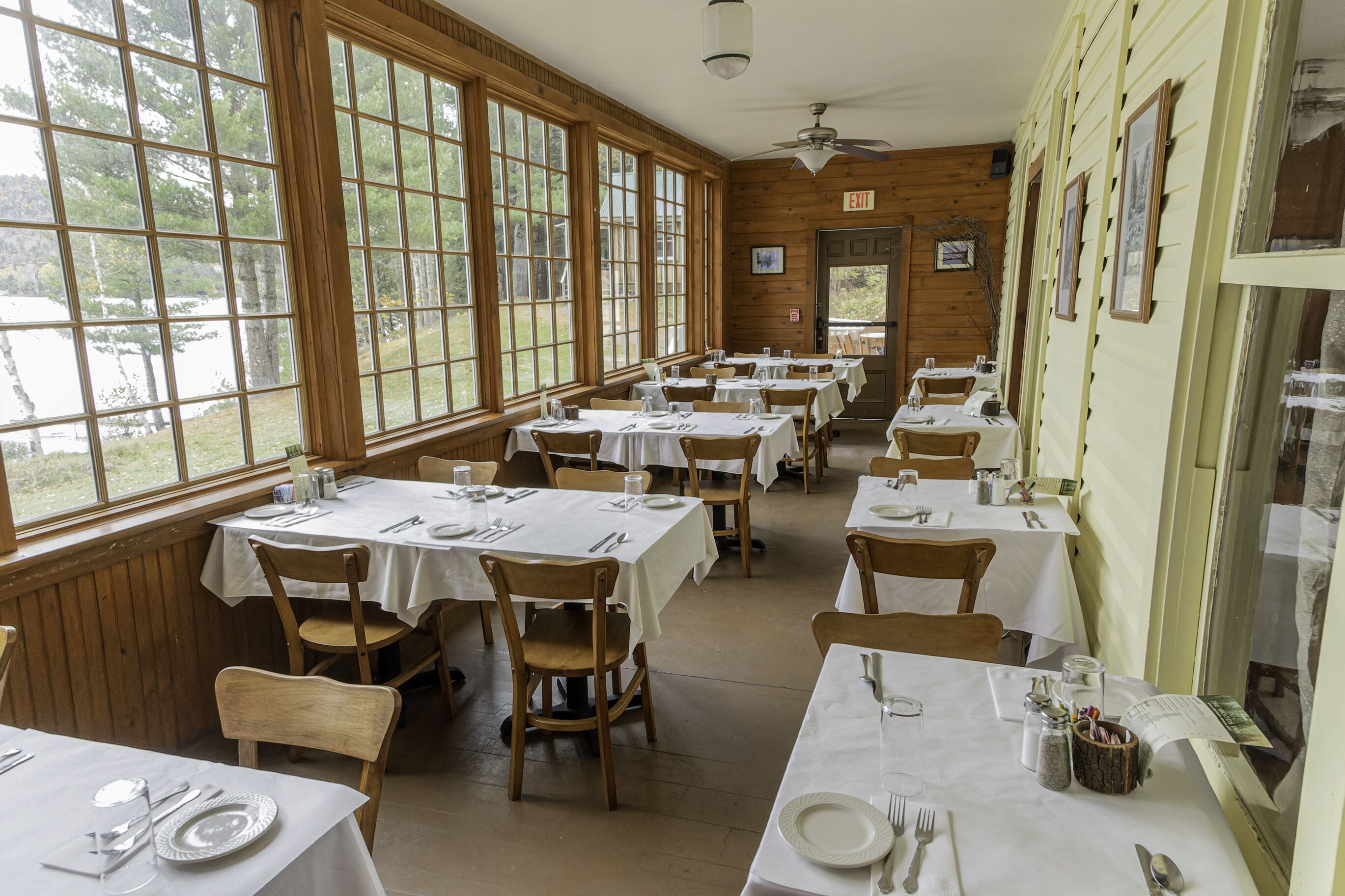 A dining area with multiple tables set for guests, large windows offering views of the outdoors, and wooden walls.