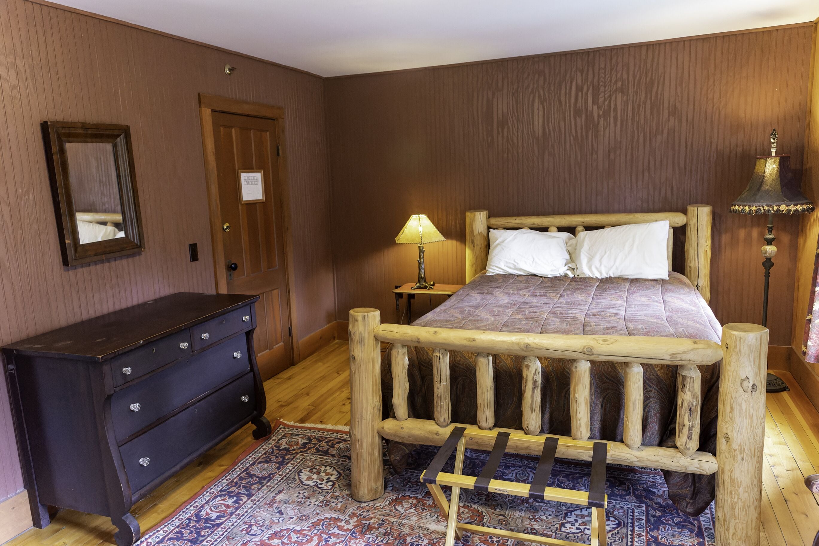 Een bed met houten frame en een beddegoed met patroon, een donkere dressoir met laden, een spiegel, een houten deur en een lamp op een nachtkastje.