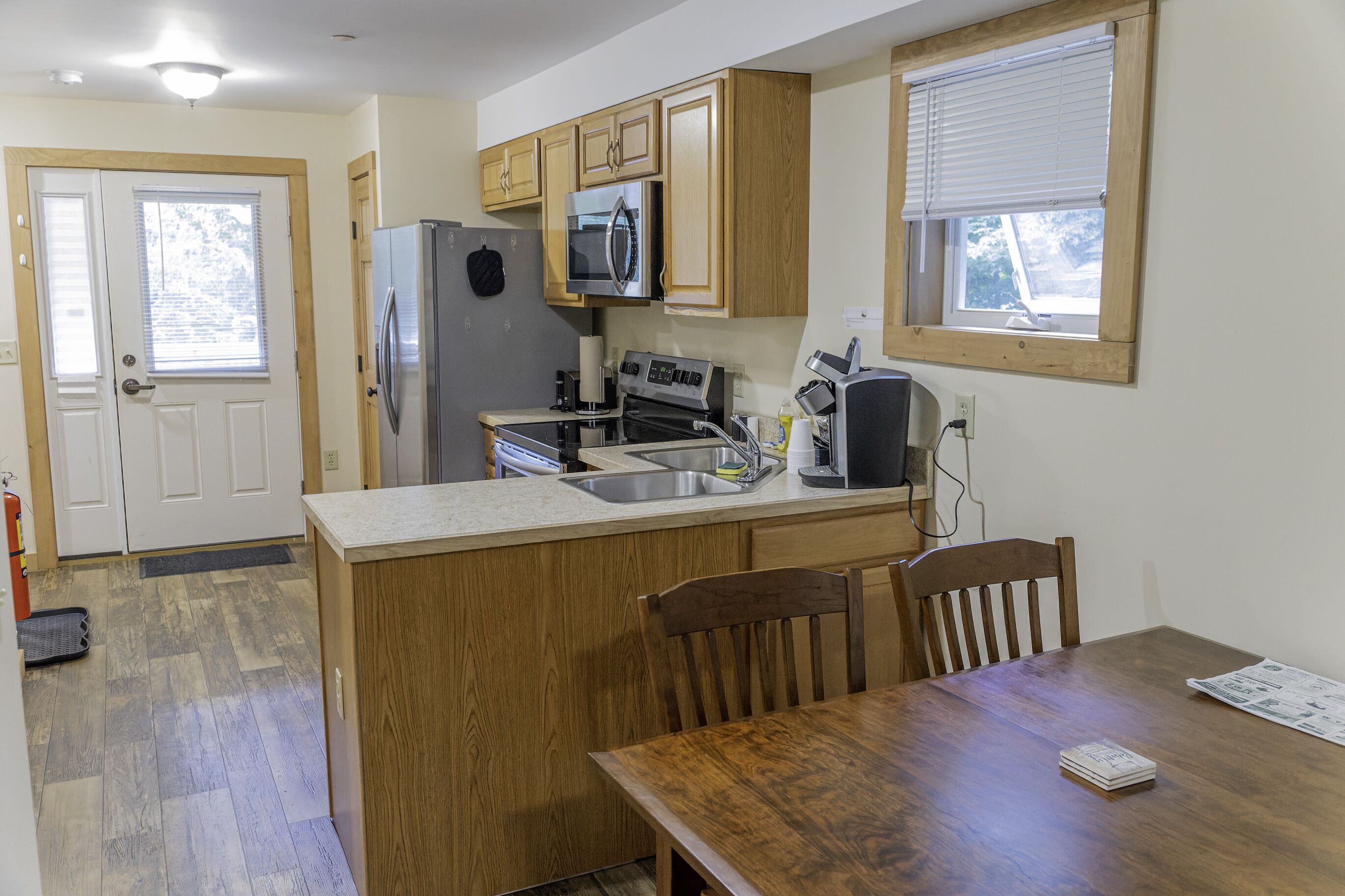 A compact kitchen with wooden cabinets, a refrigerator, a microwave, and a stove. There is a dining table with chairs and a window with blinds.
