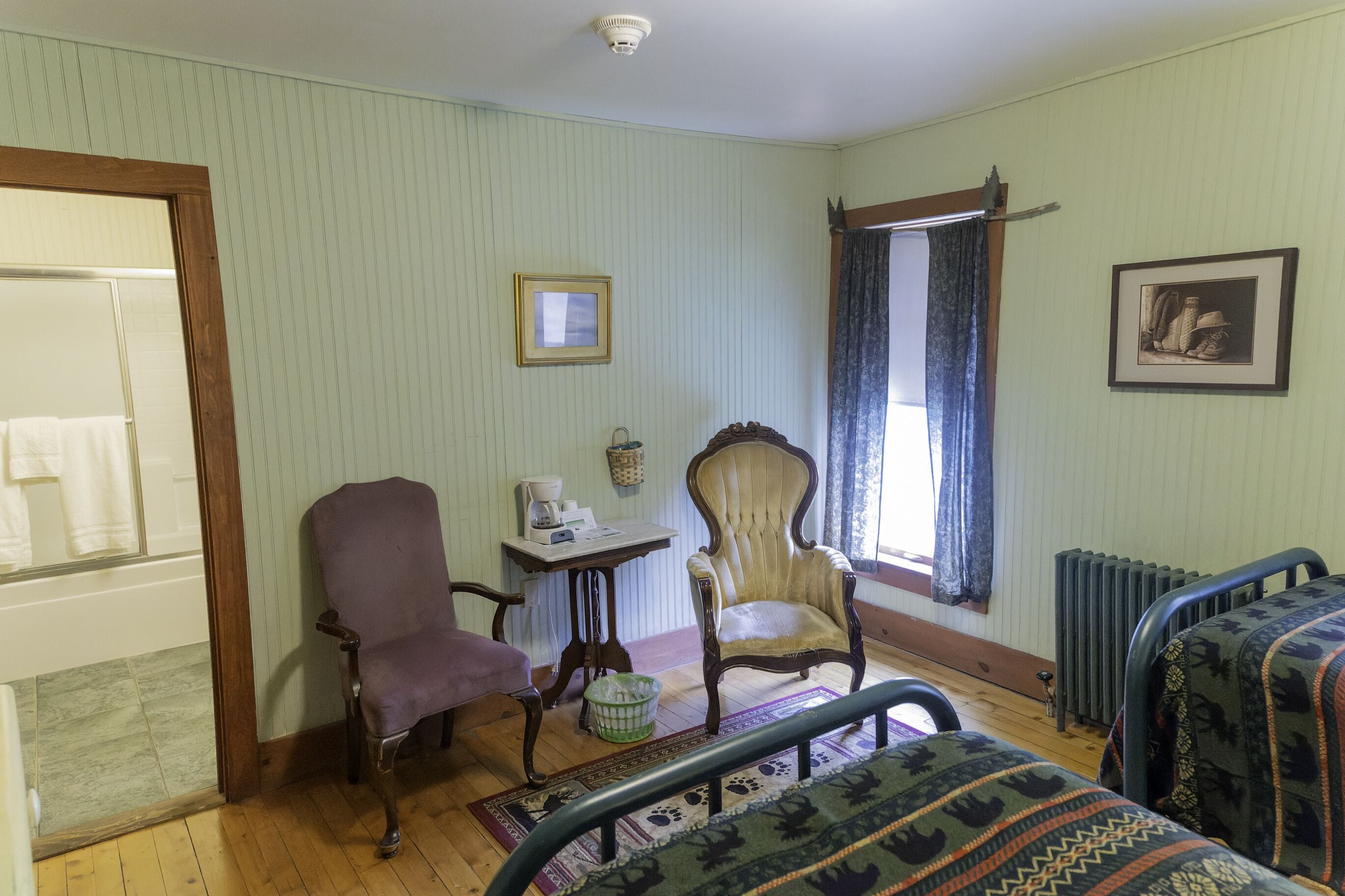 Una habitación con suelo de madera, una silla morada, una mesa de madera con una cesta, una silla con cojín, un cuadro enmarcado en la pared y un radiador.