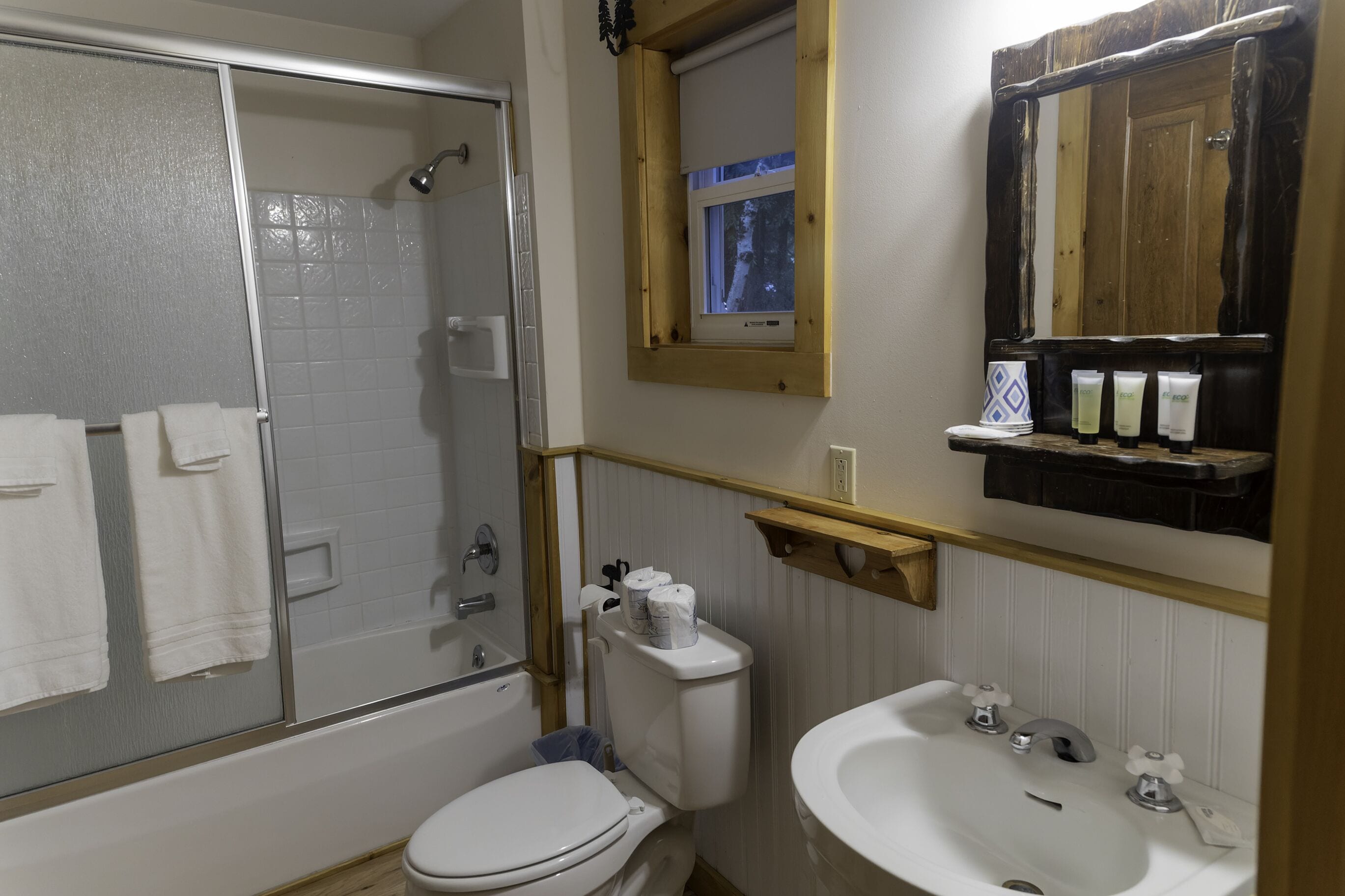 Un baño con ducha, inodoro, lavabo y una ventana sobre el espejo.